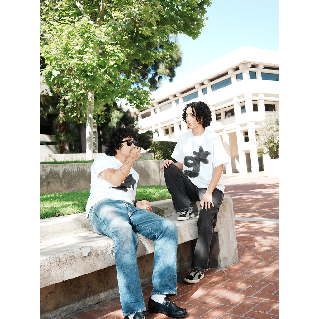 Two people sitting on a concrete bench outdoors with trees and a building in the background wearing g & star white t-shirt