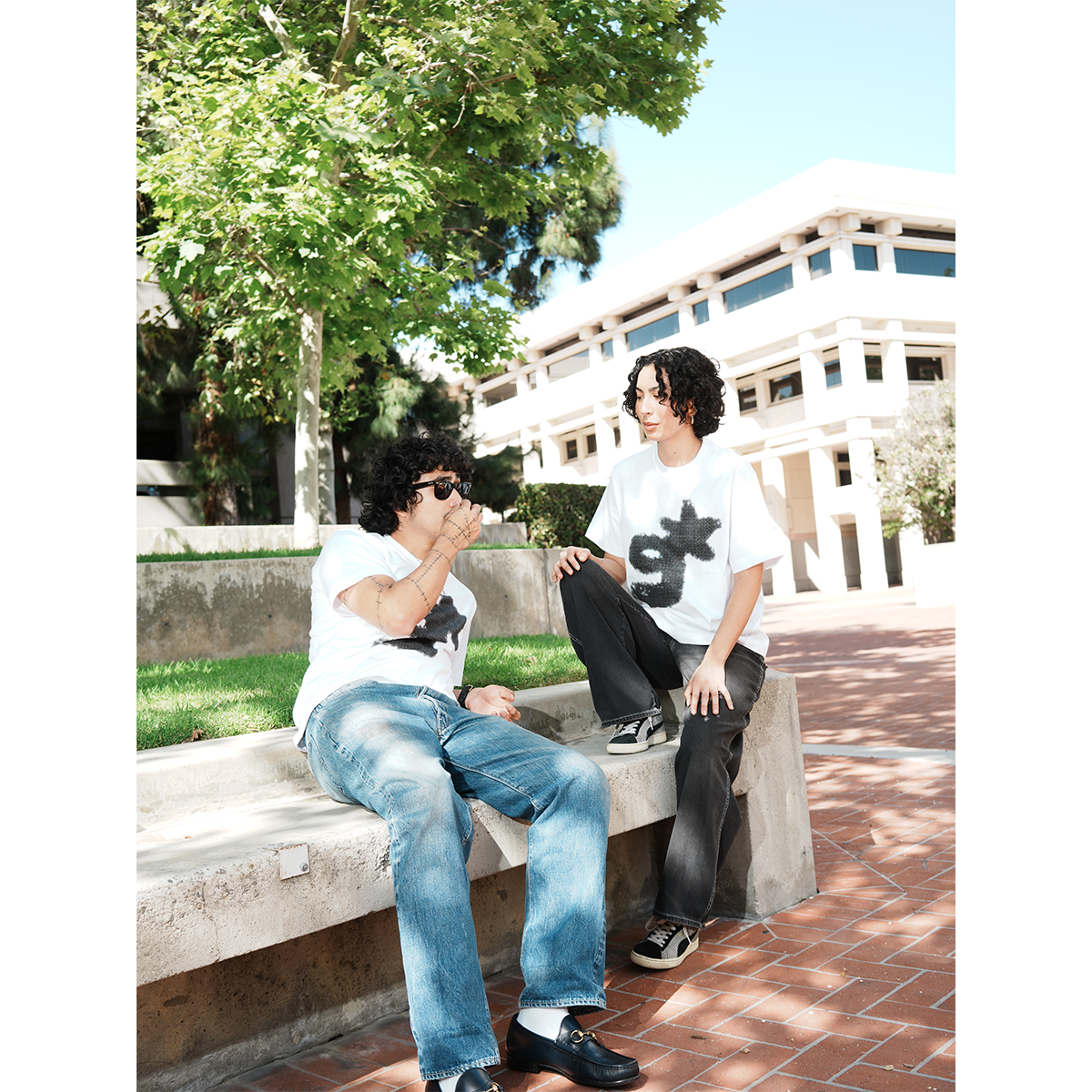 Two people sitting on a concrete bench outdoors with trees and a building in the background wearing g & star white t-shirt