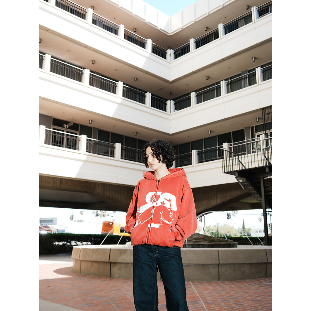 Model wearing a red hoodie with a white graphic design in an outdoor setting with hands in pocket.