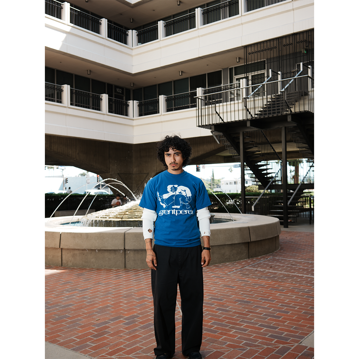 Model wearing a blue t-shirt with a graphic design standing in an outdoor setting with a fountain and building.