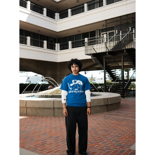 Model wearing a blue t-shirt with a graphic design standing in an outdoor setting with a fountain and building.